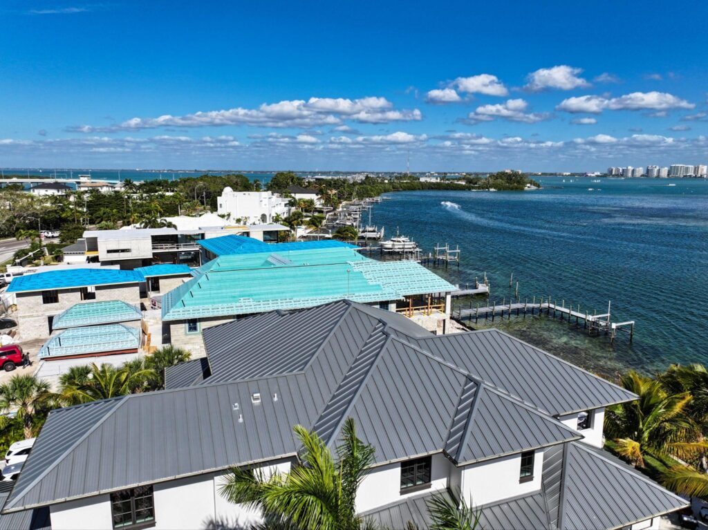 High-angle coastal perspective showing a completed dark gray roof adjacent to active construction sites, utilizing a SUNLOC Standing Seam layout by Sunshine Metal Supply.
